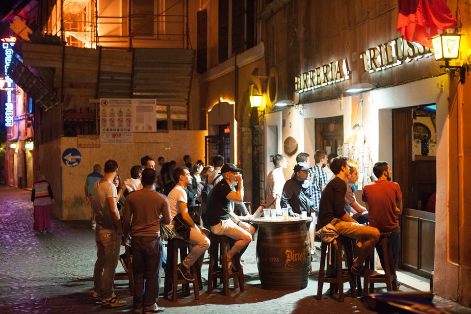 Ludzie przy stołach z beczek przed pubem piwnym Birreria Trilussa nocą na Zatybrzu w Rzymie